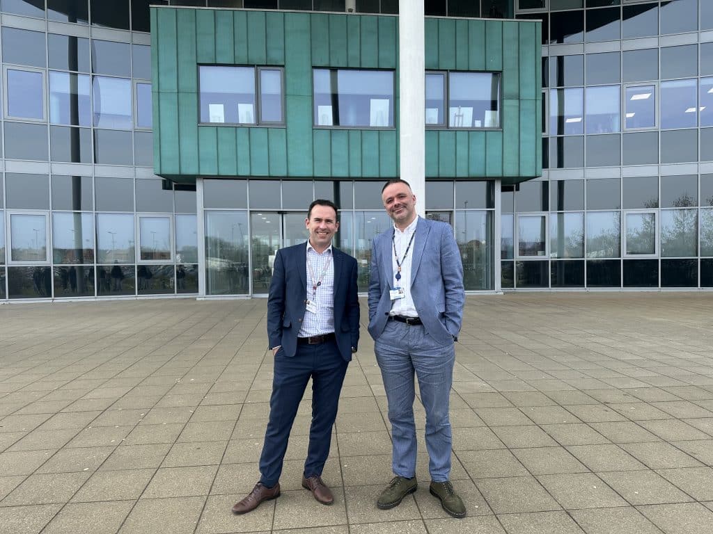 two men in suits standing outside large building with lots of windows smiling
