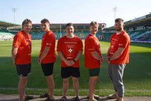5 men in rugby kit standing on football pitch, red rugby tops